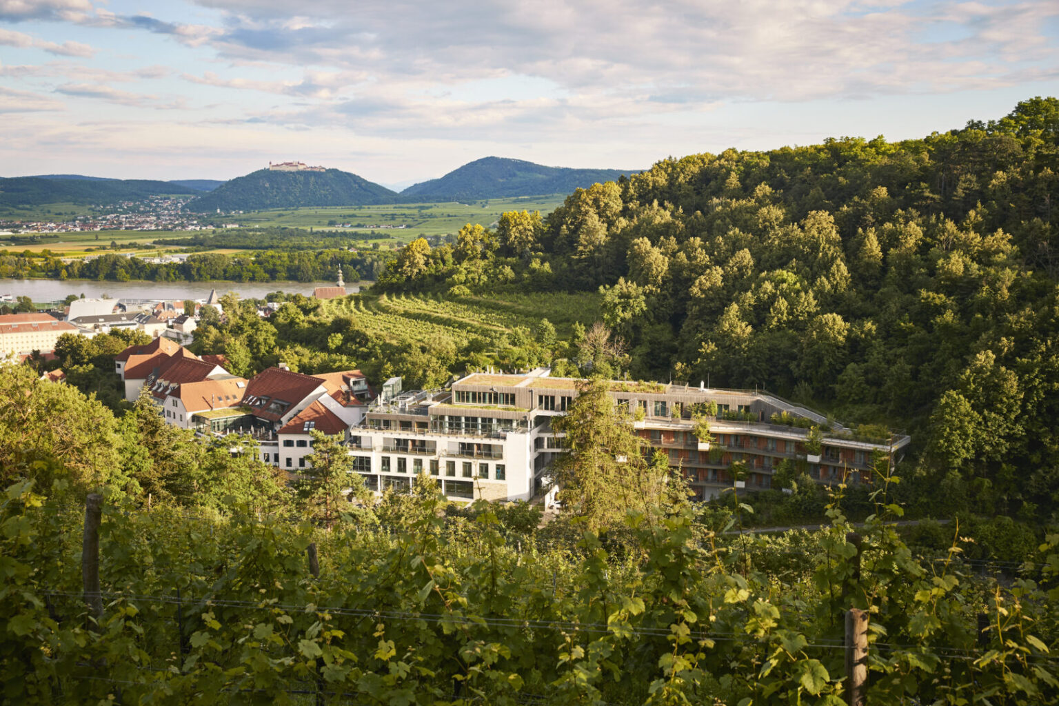 Außenansicht des Steigenberger Hotel & Spa Krems mit moderner Architektur, großen Glasfenstern und einer Terrasse mit Sitzmöglichkeiten, umgeben von grünen Hügeln.