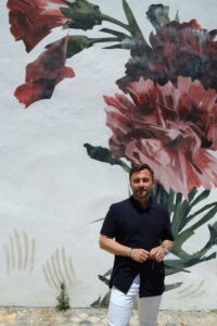 Ein Mann steht vor einer Wand, die mit bunten Blumen bemalt wurde und lächelt in die Kamera.