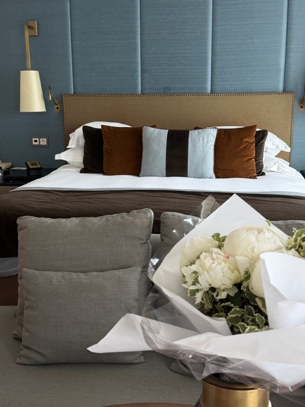 Ein Hotelzimmer mit großem Bett, auf dem viele Kissen liegen und vor dem ein Blumenstrauß steht.