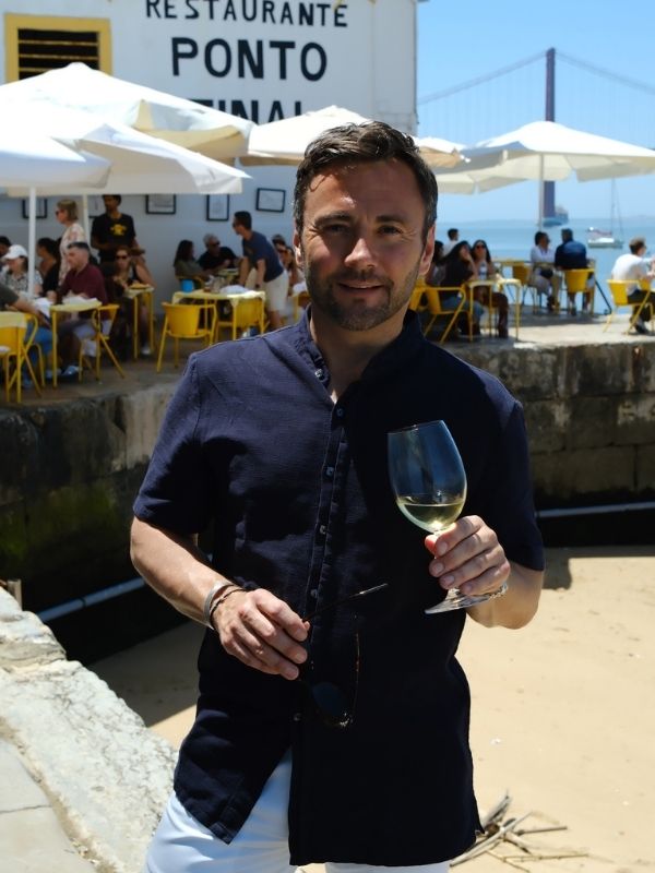 Ein Mann steht mit einem Glas Wein vor der Terrasse eines Restaurants, im Hintergrund sieht man gelbe Stühle und Tische, Menschen und eine Brücke.