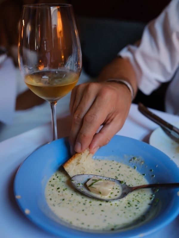 Ein Hand tunkt ein Stück Brot in eine Sauce, im Hintergrund sieht man ein Glas mit Weißwein.