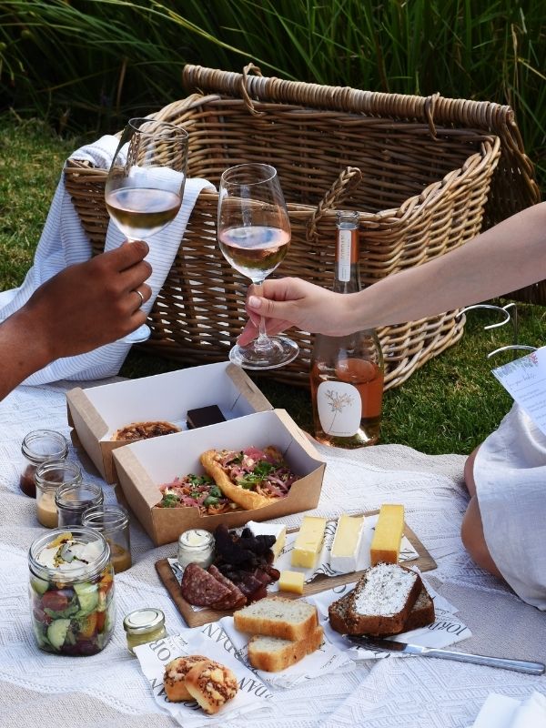 Ein Picknickkorb, verschiedene kleine Gerichte und zwei Hände mit mit Weingläsern anstoßen.